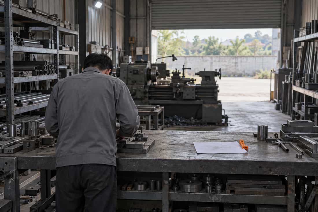 Xưởng cơ khí nhìn rộng từ cuối xưởng: người quay lưng đứng bên trái bàn thép, tờ giấy trắng bên phải có kẹp cam