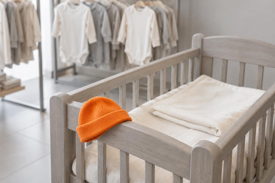 Tiệm đồ sơ sinh buổi sáng, chiếc nôi gỗ có mũ len màu cam trên mép nôi, giá treo áo trắng mờ phía sau