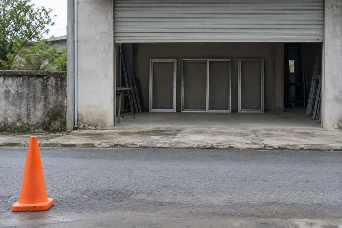 Mặt tiền xưởng cửa nhôm kính cửa cuốn kéo nửa nhìn từ bên kia đường vắng, cọc nón giao thông cam ở tiền cảnh
