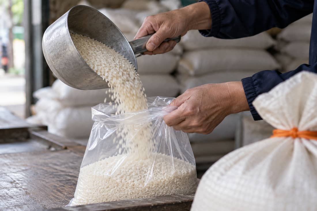 Cửa hàng gạo cận cảnh miệng túi: gạo đổ từ ca nhôm vào túi trong, bàn tay giữ túi, bao tải dây cam