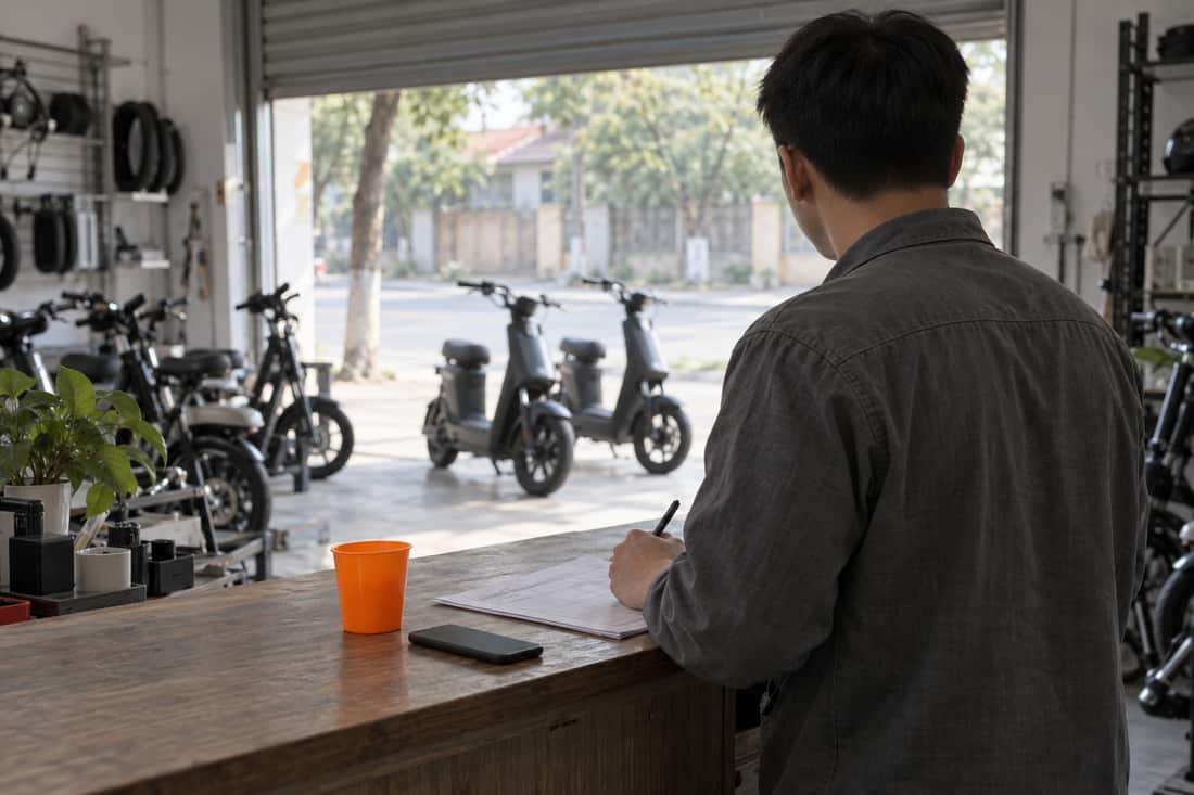 Cửa hàng xe đạp điện nhìn từ phía sau: người đứng sau quầy nhìn ra cửa cuốn, tay cầm bút, cốc nhựa cam trên quầy