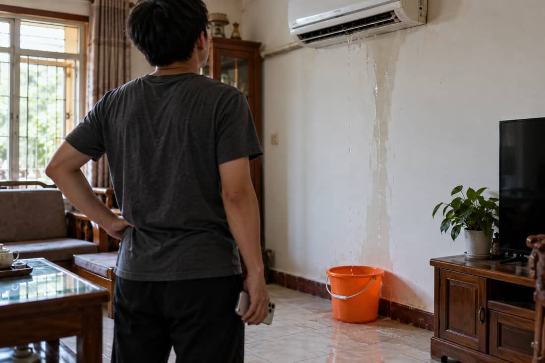 Một người đứng quay lưng nhìn chiếc điều hoà trắng đang chảy nước thành vệt xuống tường, một tay buông điện thoại thấp bên hông, dưới sàn có chiếc xô nhựa màu cam hứng nước