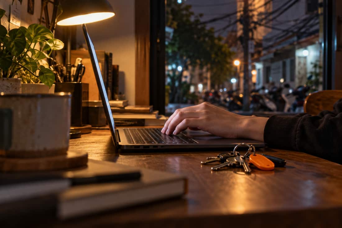 Bàn tay đặt trên bàn phím laptop, cạnh chùm chìa khoá móc cam, ngoài cửa sổ mờ là phố nhỏ có xe máy